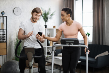 Obraz premium Young couple doing cardio on stationary bike and treadmill using smart phones to control physical parameters. Attractive female and male working out on fitness at home.