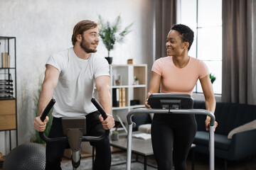 Fototapeta na wymiar Home fitness workout sporty people training on exercise machines indoors. Portrait of focused caucasian male and african female wearing sportswear using exercise bike and treadmill.