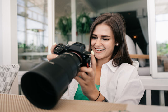 Adorable female young brunette model  hods camera watching photos on screen, smiling satisfied by shooting results. Attractive young American woman at cafe flipping through photos. 