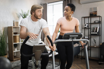 Fototapeta premium Portrait of focused caucasian male and african female wearing sportswear using exercise bike and treadmill. Home fitness workout sporty people training on exercise machines indoors
