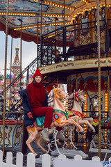 Obraz na płótnie Canvas cheerful woman in red clothes, a knitted sweater and a hat, rides a horse on a carousel on red square in Moscow on Christmas in winter.