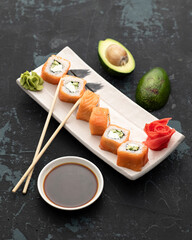 Set of Philadelphia sushi rolls on white plate with chopsticks and soy sauce. Served with ginger and wasabi. Decorated with cut avocado on table. Dark background. View from above. 