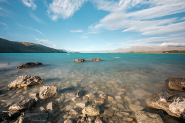 Blue skies over the turquoise blue water of Lake Tekapo in New Zealand with snow capped mountains in the distance and the rocky shoreline in the foreground