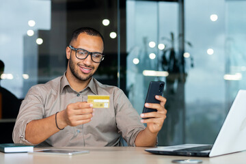 Successful hispanic businessman inside office, man in shirt smiling and happy holding bank credit card and phone, making money transfer and online shopping.