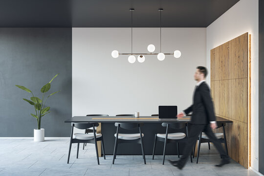 Side View Blurred Businessman Walking By Dark Big Meeting Table With Modern Laptop In Stylish Conference Room Interior Design With Lamps On Top And Light Wall Background With Wooden Part