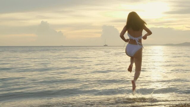 Back view, Young beautiful woman wear bikini swimsuit running to tropical beach and jumping raise hand freedom, Happy Asian female having fun and jump in holiday outside sunset, Summer travel vacation