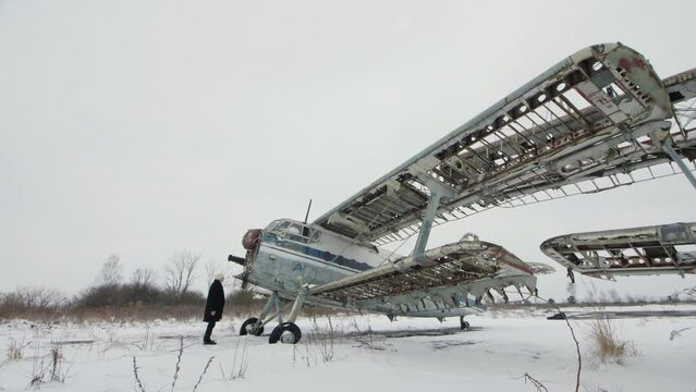 A man in black clothes stands in front of an abandoned broken Soviet aircraft. He turns and spreads his arms like wings. Winter landscape, falling snow.