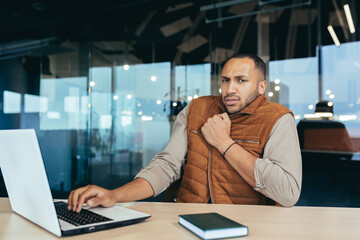 African american man freezing in office, no heating in workplace, man in outerwear sitting at desk using laptop at work.