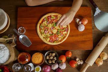 Home made Italian pizza served on the wooden table from top view. Woman decorating pizza toping with some basil leaf and seasoning.