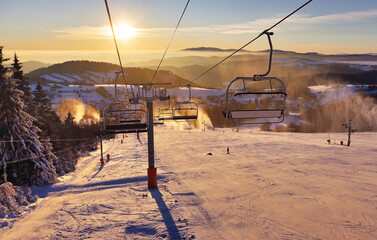 Ski lift empty ropeway on hilghland alpine mountain winter resort on bright sunny evening . Ski chairlift cable way with people enjoy skiing and snowboarding. Sunset sky backlit shining on background