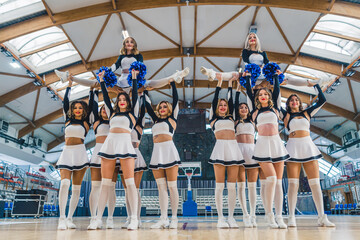 Two cheerleading teams in white and navy blue uniforms doing a gymnastic figures while holding two...