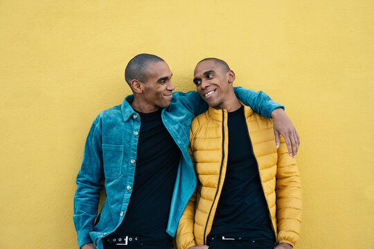 African American adult identical twin brothers hugging and smiling, standing over yellow wall 