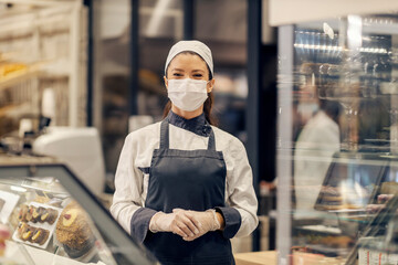Fototapeta na wymiar A bakery department worker is standing at bakery with face mask on face and looking at the camera at supermarket.