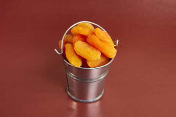 A small metal bucket filled with dried apricots on a brown background. Dried apricots as an ingredient.