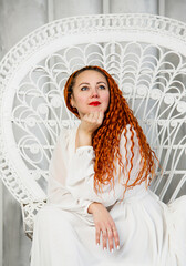 beautiful red-head girl in white dress with long curly hair  sitting in white chair in studio 