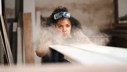 Female multiracial carpenter working in woodshop blowing sawdust from plank. Afro woman with...