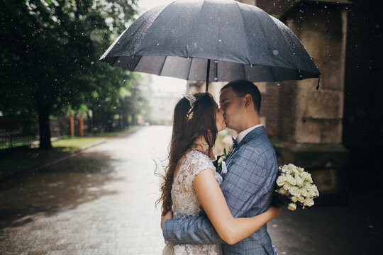 Stylish Bride With Bouquet And Groom Kissing Under Umbrella On Background Of Old Church In Rain. Provence Wedding. Beautiful Wedding Couple Embracing Under Black Umbrella In Rainy Street. Romantic