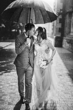 Beautiful wedding couple embracing together under umbrella in sunny rainy street. Stylish bride and groom walking and kissing on background of old church in rain. Black and white photo
