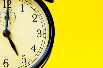 Closeup of a vintage analog black clock on a yellow background displaying 5 o'clock