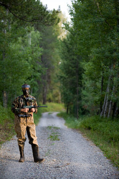 A man dressed in paintball garb poses on a forest trail.
