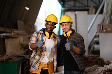 Smart African American and white carpenter showing thumb up at carpentry shop, wearing yellow helmet and safety goggles to protect dust and accident while working at a machine. Teamwork collaboration.