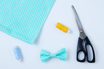Turquoise bow tie with white polka dots, scissors, fabric and sewing threads on a white background.