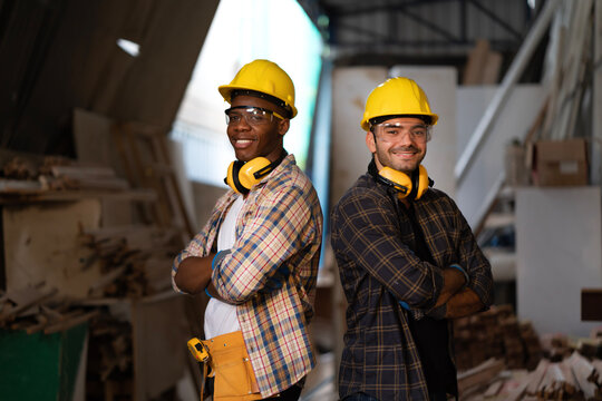 Portrait of young carpenter team with yellow helmets, safety goggles and yellow ear muffs standing with smiling faces and arms crossed in carpentry shop. Happy builder worker making handmade furniture