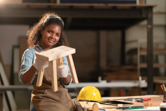 African American young carpenter woman feeling happy and smiling face successfully makes a small chair in hands in a carpentry shop. Professional handmade design furniture on customer order. 