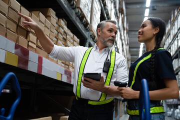 Elderly warehouse manager holding tablet in hand with walkie-talkie, pointing barcode on boxes and coaching junior worker to scan and update online inventory. Manufacturing factory supplier.
