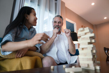 Hispanic husband and Asian wife playing board game in living room, building wooden tower together at home. Having fun together, family relationship activities of recreation.