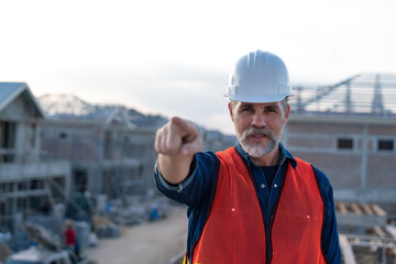 senior engineer wearing white helmet, pointing his finger in the air with home construction background at real estate project. Professional elderly civil engineering checking project plan.