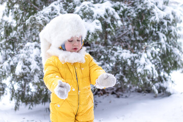 Fototapeta na wymiar child playing with snow. portrait of little girl in yellow jumpsuit and fluffy white hat brushing snow off mittens on snowy winter day in the park