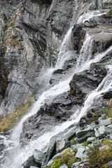 Foaming streams of alpine waterfall shot in contrast and with high sharpness against background of granite dark rocks overgrown with sparse green vegetation and herbs, Aosta valley, Italy