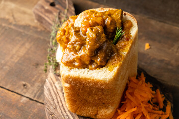Bunny chow curry with meat and vegetables served in white bread and carrot salad on a wooden background, close-up. - obrazy, fototapety, plakaty