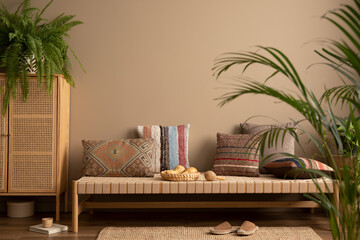 Ethno composition of living room interior with couch, patterned pillows, rattan sideboard, braided rug, fern, plants in flowerpots, brown slippers, cup and personal accessories. Home decor. Template.