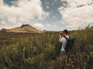 Caucasian young man with dark hair and bag photographing landscape with cacti and mountains in...