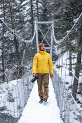 Young caucasian man walking on a hanging bridge on a snowy day holding a camera in winter clothes.
