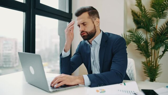 Stressed worried upset tired caucasian businessman, corporate director, financial manager, sits in the office at workplace, nervous of deadline a project, closed his eyes, holding hand on head