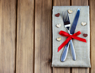 Romantic dinner concept. Top view of table appointments: napkin, silver fork and knife decorated red bow and hearts on wooden background