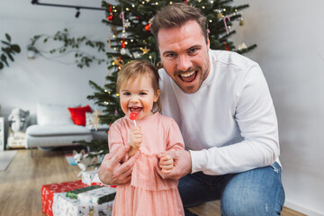 Portrait of smiling cheerful dad with is baby girl in a pretty pink dress standing in front of the Christmas tree