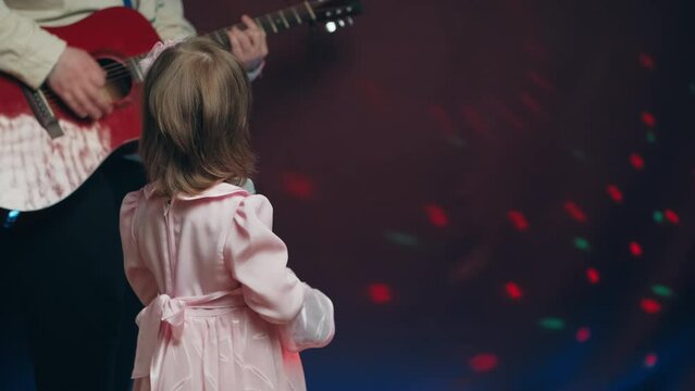Little girl on stage in vintage dress, she sings into microphone and dances, her father plays acoustic guitar. Color music is shining. Performance on stage. Early development of children