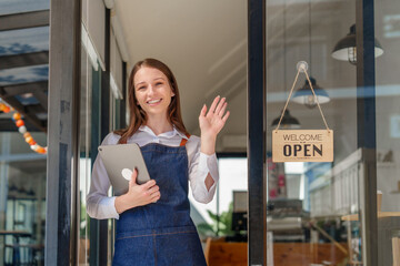 Young friendly female pretty caucasian coffee shop woman owner in apron with open sign, small business owners smiling and service take away orders from food delivery application on tablet.