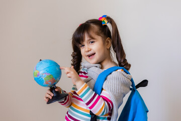 a happy little preschool girl with a backpack and a globe on a light background, space for text. Preschool education, traveling with children