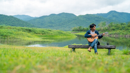 Young Asian man drinking coffee and playing guitar by the lake in the morning. Handsome guy relax and enjoy outdoor lifestyle hiking and camping in forest mountain on summer holiday travel vacation. - Powered by Adobe