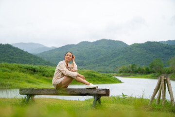 Naklejka na ściany i meble Young Asian woman enjoy outdoor lifestyle hiking and camping in forest mountain while travel on summer holiday vacation trip. Attractive girl traveler resting on outdoor chair by the lake in morning.