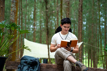 Asian man relax and enjoy outdoor lifestyle camping in forest mountain on summer holiday travel vacation. Handsome guy resting on outdoor chair and reading a book in pine tree forest in the morning