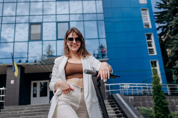 Beautiful young woman in sunglasses and white suit standing with her electric scooter near modern building and looking at camera