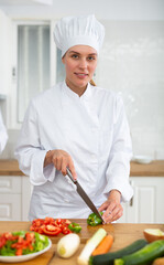 Cheerful female chef in white uniform preparing vegetable dish in private kitchen