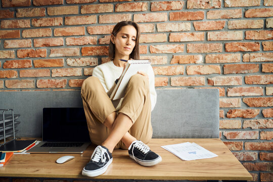 Picture of young businesswoman sitting on desk writing notes in notebook in her office
