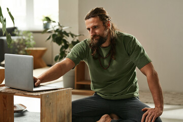 Young man sitting on the floor in the room and using laptop to do yoga online